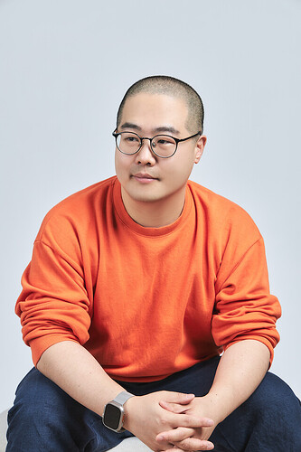 A person wearing glasses and an orange sweater with rolled-up sleeves is sitting with their hands clasped. (Captioned by AI)