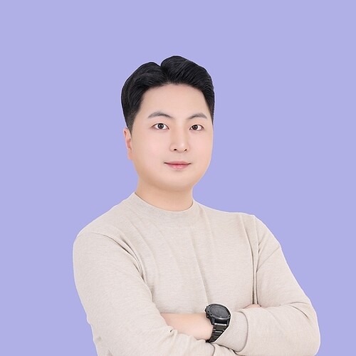 The image shows a person with short black hair and a light-colored shirt posing with arms crossed against a light purple background. (Captioned by AI)