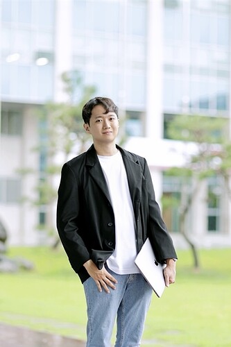 A man is standing outdoors holding a laptop, dressed in a black blazer over a white shirt and blue jeans. (Captioned by AI)